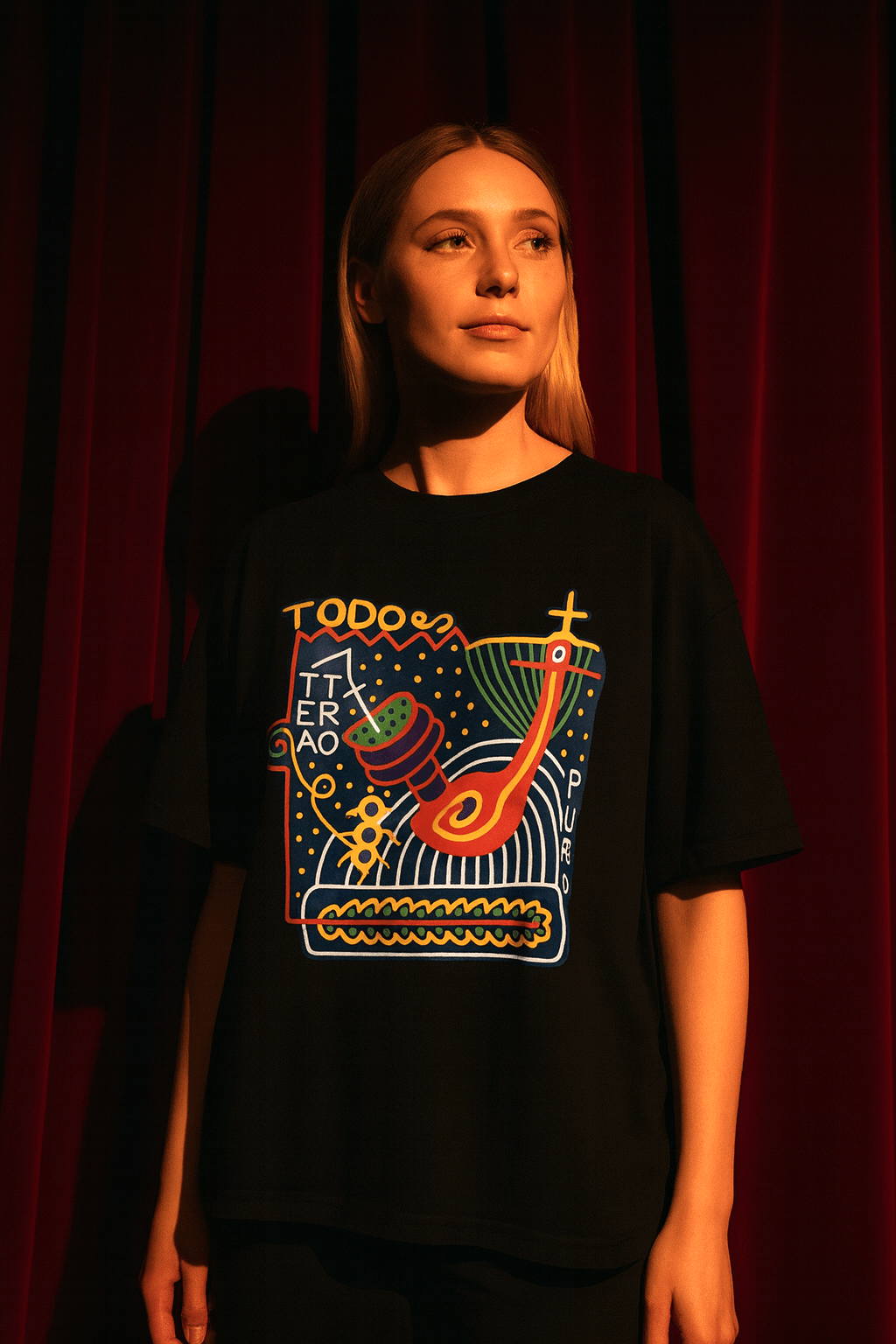 Model wearing TODO ES UN TEATRO Shirt with vibrant graphic design against red curtain backdrop.